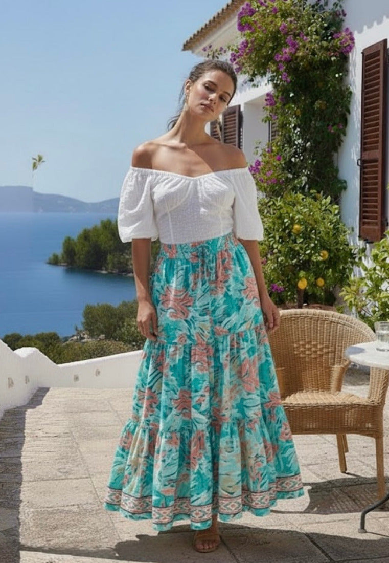 Woman in a white top and floral skirt standing on a sunlit patio with a scenic view.