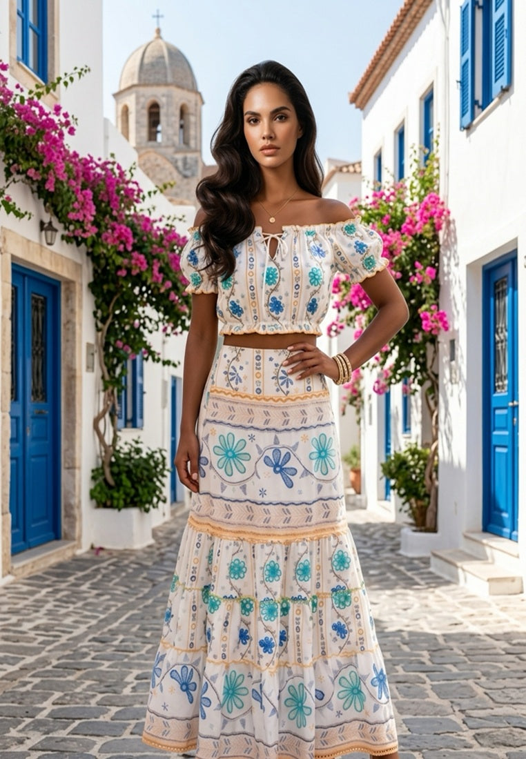 Woman wearing this Santorini Floral co-ord set from Coral Secretstanding in a picturesque alleyway with blue doors and pink flowers.