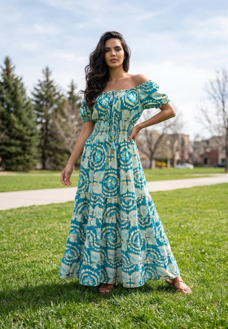 A woman stands outdoors on a grassy lawn wearing a Coral Secret flowing off-the-shoulder maxi dress with a turquoise and cream circular batik-style print.