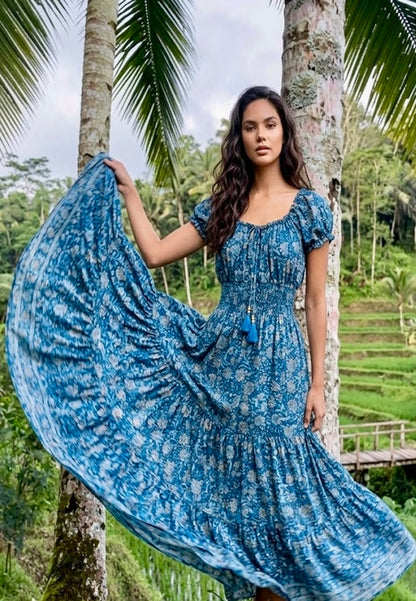 Woman Off Shoulder Silk Maxi Dress from Coral Secret standing in a tropical setting with palm trees.